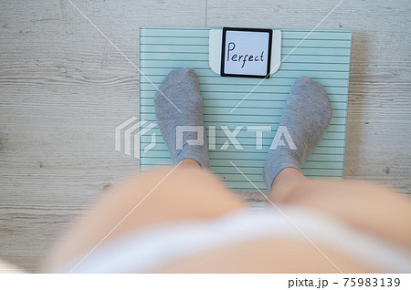 Faceless woman measures weight on the floor scales. Top view of women's feet in gray socks on the scales with the word perfect. The inscription on the screen is perfect. There is no excess fat. 75983139