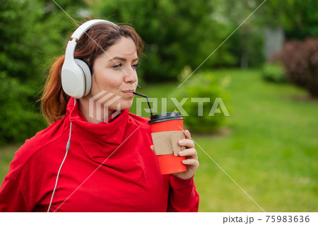 Red-haired woman listens to music on headphones and drinks coffee with a straw in the park. A girl enjoys a walk on a sunny summer day and holds a takeaway cup. earphones connected to a smartphone. 75983636