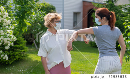 Two masked women greet their elbows in the park. An elderly woman and her daughter maintain social distance during the coronavirus epidemic. A new way of shaking hands. Apple trees are blooming. 75983638