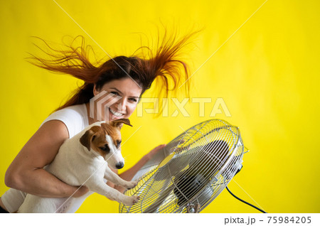 Beautiful caucasian woman and a dog are cooling off by an electric fan. A girl with her pet Jack Russell Terrier freshen up at the air conditioner. Red hair develops under the blow of cold air. 75984205