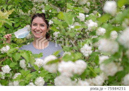 Happy red-haired woman takes off a medical mask while standing next to a blossoming apple tree in a park. Ending seasonal allergic rhinitis. The girl was cured of an allergy to pollen. 75984251