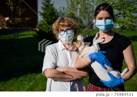 A woman holds a puppy and her elderly mother is dressed in medical masks to protect against the virus. Retired woman and her adult daughter. Dog walking in the park during the coronavirus epidemic. 75984531