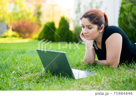 A woman lies on her stomach on the lawn at the cottage and uses on a laptop. A focused female freelancer works remotely outdoors. The girl is studying on the computer. A woman lies on her stomach on the lawn at the cottage and uses on a laptop. A focused female freelancer works remotely outdoors. The girl is studying on the computer. 75984846