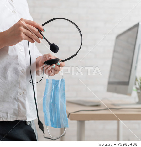 A faceless female employee holds a headset and a face mask. Woman with headphones and a medical mask in her hands 75985488