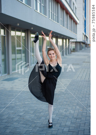 Ballerina in tutu posing against a residential building. Beautiful young woman in a black dress and pointe shoes jumping demonstrates stretching. Ballerina is standing in the splits. Outside. Ballerina in tutu posing against a residential building. Beautiful young woman in a black dress and pointe shoes jumping demonstrates stretching. Ballerina is standing in the splits. Outside. 75985584