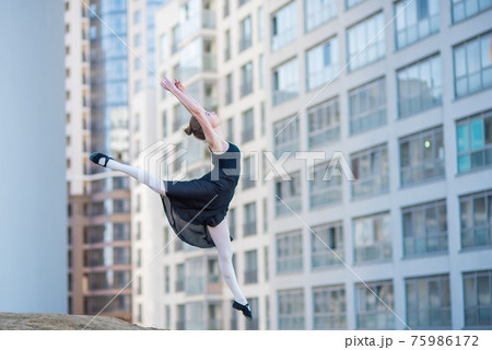 Ballerina in tutu posing against a residential building. Beautiful young woman in black dress and pointe shoes jumping with incredible flexibility. Ballerina performs elegant jumping with deflection. 75986172