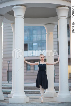 Ballerina in a black dress is a beautiful pose. A beautiful young woman in a black bodysuit is dancing in an elegant classical dance on a background of columns. 75986418