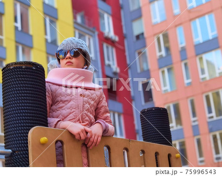 The girl 9 years old on the playground. The child is dressed in warm demi-season clothes a pink jacket and a grey hat. 75996543