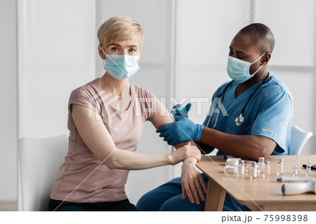 Adult african american lady sitting at doctor's office and getting modern Covid 19 shot injection in clinic Adult african american lady sitting at doctor's office and getting modern Covid 19 shot injection in clinic 75998398