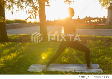 Cute young pregnant woman in black tracksuit makes heroe pose standing barefoot on rug on green lawn on sunny summer day. Childbirth preparation and nervous calming concept 76002449