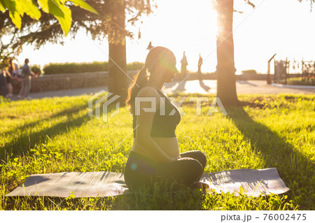 Peaceful young positive pregnant woman in gymnastic suit does yoga and meditate sitting on mat on green grass on sunny warm summer day. Concept of preparation for childbirth and positive attitude Peaceful young positive pregnant woman in gymnastic suit does yoga and meditate sitting on mat on green grass on sunny warm summer day. Concept of preparation for childbirth and positive attitude 76002475