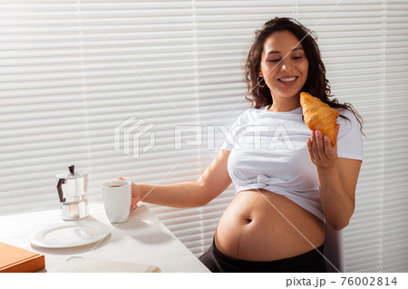 Joyful pregnant young beautiful woman eating croissant and drinking tea during morning breakfast. Concept of pleasant morning and expectation of meeting baby. Copyspace Joyful pregnant young beautiful woman eating croissant and drinking tea during morning breakfast. Concept of pleasant morning and expectation of meeting baby. Copyspace 76002814