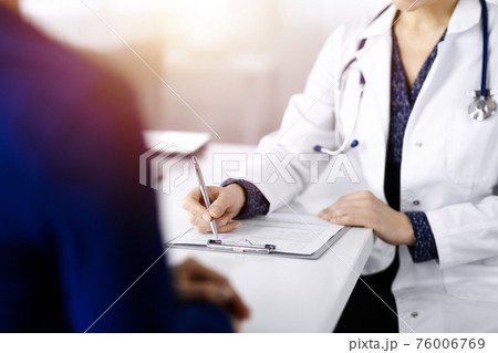Unknown woman-doctor is writing some medical recommendations to her patient, while they are sitting together at the desk in the sunny cabinet in a clinic. Physician is using a clipboard, close-up Unknown woman-doctor is writing some medical recommendations to her patient, while they are sitting together at the desk in the sunny cabinet in a clinic. Physician is using a clipboard, close-up 76006769