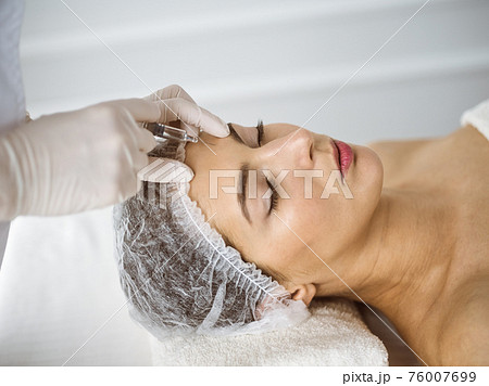 Beautician doing beauty procedure with syringe to face of young brunette woman in sunny clinic. Cosmetic medicine and surgery, beauty injections 76007699