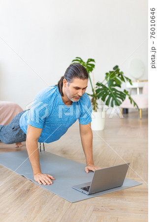 Vertical photo of a middle-aged man doing push-ups in front of laptop. He uses an online tutorial from the internet to monitor the workout. Vertical photo of a middle-aged man doing push-ups in front of laptop. He uses an online tutorial from the internet to monitor the workout. 76010646