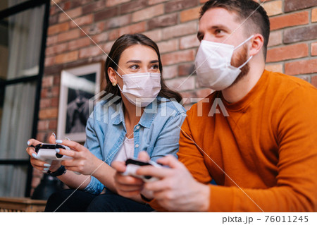 Close-up portrait of happy young couple wearing medical protective masks playing video games on console 76011245