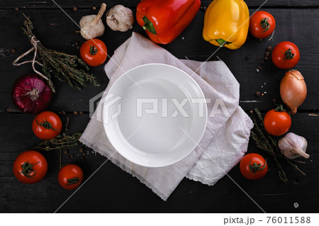 White clean plate on a napkin, bell peppers, tomatoes, garlic and onions are laying on a dark wooden table. Contrast bright colored flatlay. Top view. 76011588