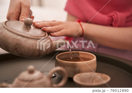Woman sitting at the floor and pouring tea at the cups for her tea ceremony 76012890