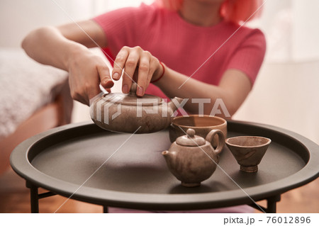 Woman with pink hair sitting at the room and pouring tea at the cups 76012896