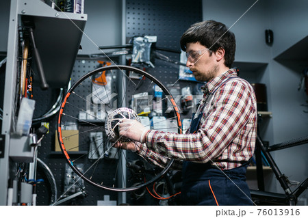 Technical expertise taking care bicycle shop. Handsome young mechanic fixing cycle wheel in workshop. Handsome repairman in workwear serving mountain bicycle. Male engineer adjusting velocipede 76013916