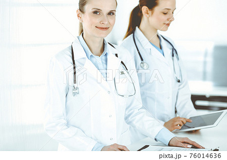 Woman-doctor cheerful smiling at work with colleague and patient at background. Portrait of middle aged female physician filling up medical documents or prescription while standing in hospital Woman-doctor cheerful smiling at work with colleague and patient at background. Portrait of middle aged female physician filling up medical documents or prescription while standing in hospital 76014636