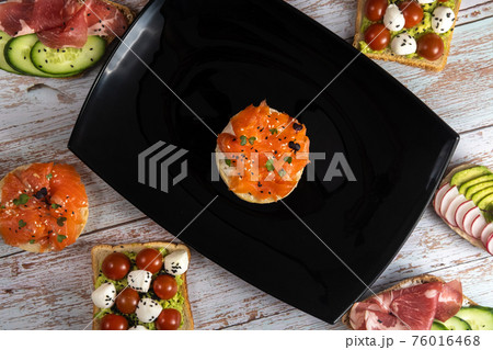 A red fish sandwich is on a black plate and there are many other sandwiches around A red fish sandwich is on a black plate and there are many other sandwiches around 76016468