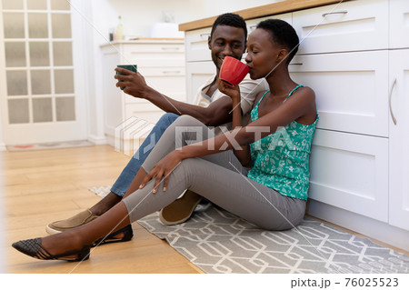 African american couple sitting on floor in kitchen holding mugs 76025523