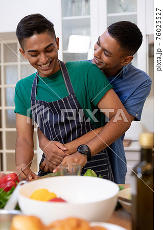 Diverse gay male couple spending time in kitchen embracing and smiling 76025527