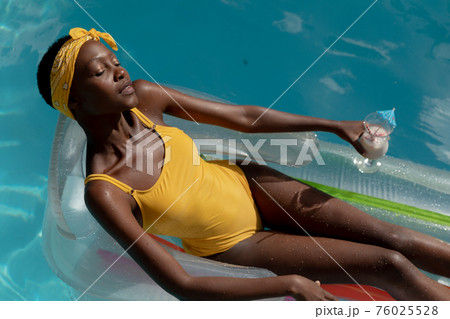 African american woman in swimming pool sunbathing on inflatable and holding drink 76025528