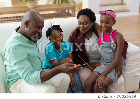 Happy african american grandfather and grandmother on couch with grandchildren looking at smartphone 76025854