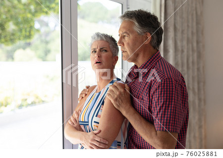 Concerned senior caucasian couple standing looking out of window embracing Concerned senior caucasian couple standing looking out of window embracing 76025881