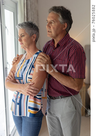 Concerned senior caucasian couple standing looking out of window embracing Concerned senior caucasian couple standing looking out of window embracing 76025882
