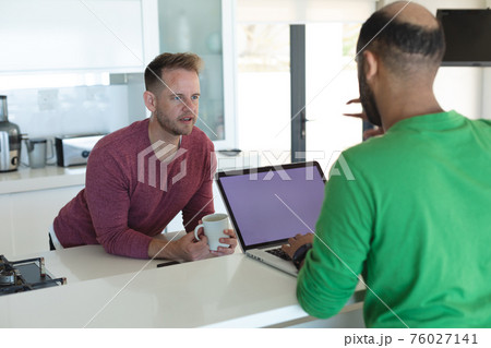 Multi ethnic gay male couple drinking coffee using laptop with copy space blank screen in kitchen 76027141