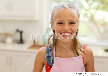 Portrait of smiling caucasian girl wearing apron standing in kitchen 76027200
