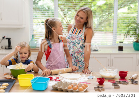 Happy caucasian mother in kitchen with daughter and son, wearing aprons baking cookies together 76027201