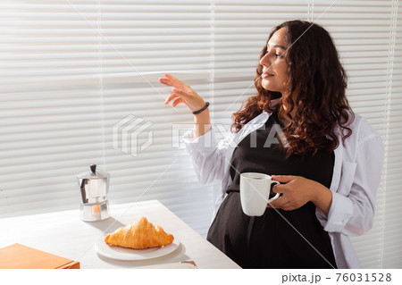 Joyful pregnant young beautiful woman looks through blinds during her morning breakfast with coffee and croissants. Concept of good morning and waiting for meeting with a baby. Copyspace 76031528