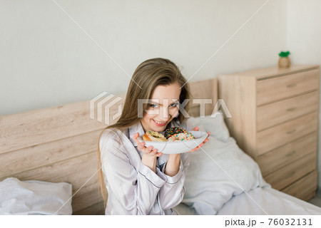 Young woman in white towel and robe in kitchen during quarantine. Hold donut on plate. 76032131