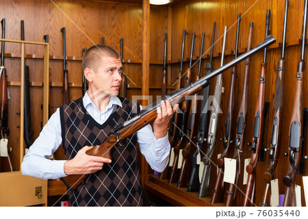 Man examining hunting shotgun before buying in gun store 76035440