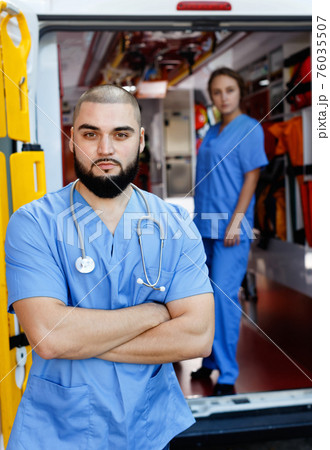 Two confident paramedicals in uniform posing near ambulance car Two confident paramedicals in uniform posing near ambulance car 76035507