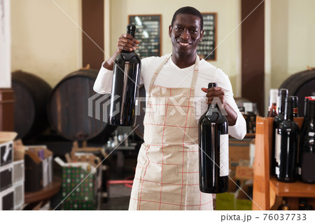 afro male seller of wine shop offering wine bottle to buy 76037733