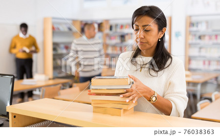 Latin american woman returning books to librarian 76038363