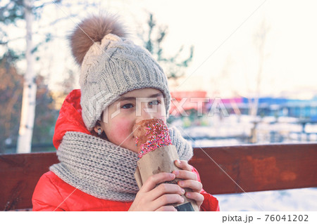 Young cute girl in bright winter clothing is eating tasty colorful pie. Happy childhood, outdoor activities. 76041202