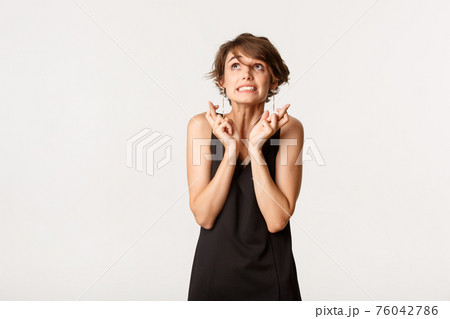 Image of hopeful woman in black dress looking up, cross fingers good luck and making wish, standing over white background 76042786