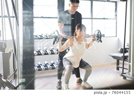 トレーナーの補助付きでバーベルスクワットをするアジア人女性 76044155