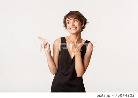 Image of pretty elegant woman smiling and pointing fingers left, standing over white background 76044302