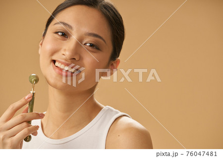 Portrait of joyful young woman smiling at camera, using jade roller for massaging her face, posing isolated over beige background Portrait of joyful young woman smiling at camera, using jade roller for massaging her face, posing isolated over beige background 76047481