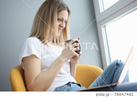 Woman holds cup of coffee and looks into laptop Woman holds cup of coffee and looks into laptop 76047482