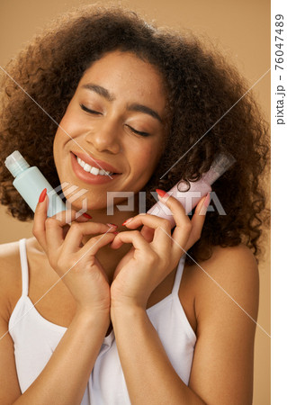 Lovely young mixed race woman looking pleased, standing with eyes closed and holding two bottles with beauty product isolated over beige background 76047489