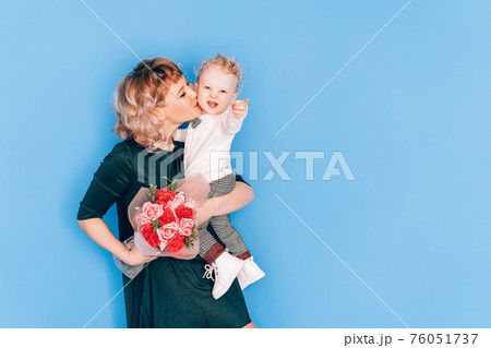 Woman in dress holding little boy in her arms and holding beautiful bouquet of flowers in against blue background. Holiday gift 76051737