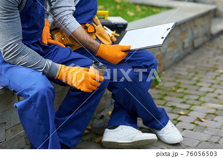 Cropped shot of two young workmen in uniform sitting outdoors having coffee while working on construction project Cropped shot of two young workmen in uniform sitting outdoors having coffee while working on construction project 76056503